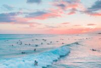 Surfers enjoying waves at a colorful beach sunset, surrounded by pastel skies.