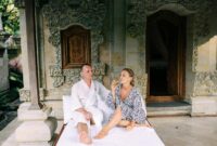 A couple in robes enjoy a relaxing moment outside a Balinese villa with ornate carvings.